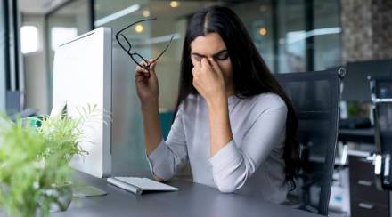 Portrait of depressed young Latin-American businesswoman sitting at computer in office holding glasses and rubbing eyes. Overworking concept