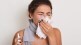 Image of sick allergic woman blowing nose, having flu or cold, sneezing into tissue, posing with closed eyes isolated on white studio background, holding nasal spray in hand.