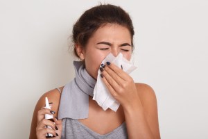 Image of ill allergic woman blowing running nose, having got flu or catch cold, sneezing in handkerchief, posing with closed eyes isolated on white studio background, holding nasal spray in hand.