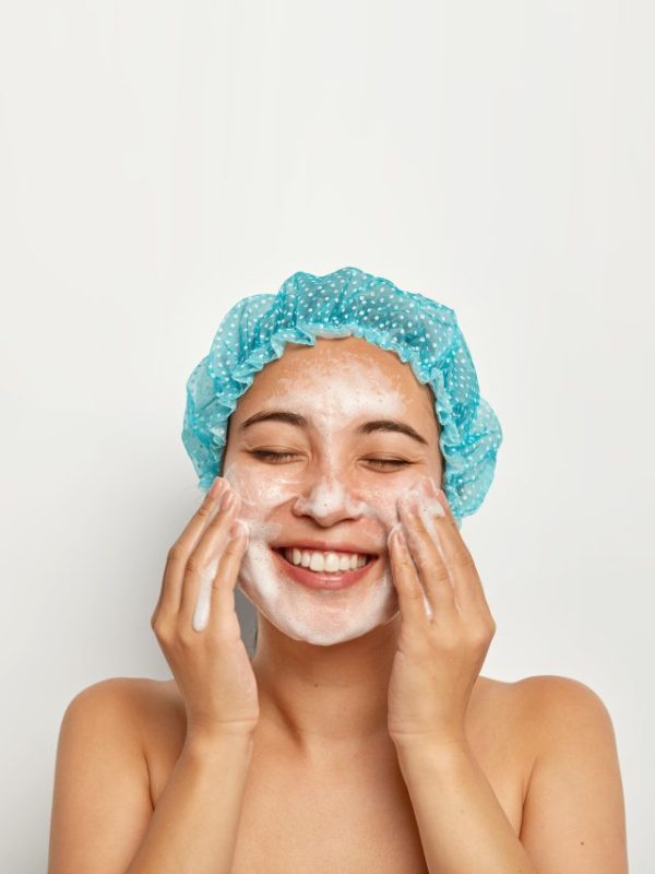 Flawless and purity of skin. Vertical image of pretty woman washes face, enjoys cold water, has foam on skin, smiles joyfully, keeps eyes closed, takes care of personal hygiene. Wellness concept