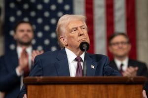 Donald Trump speaking at rally with US flag and trade charts in background