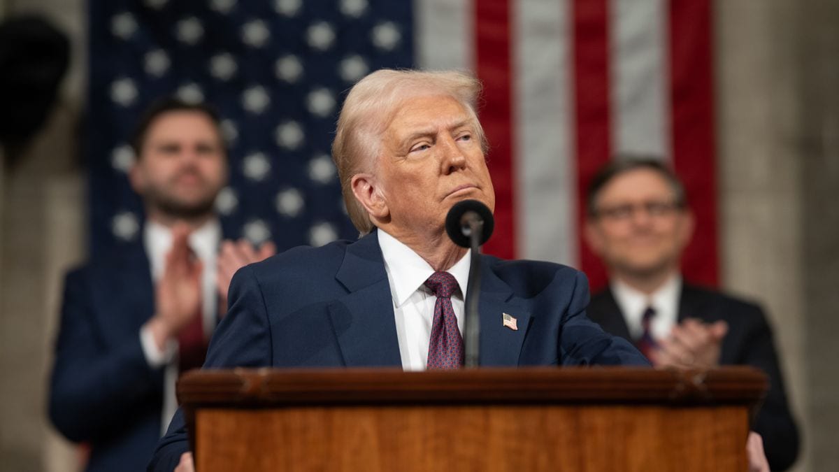 Donald Trump speaking at rally with US flag and trade charts in background